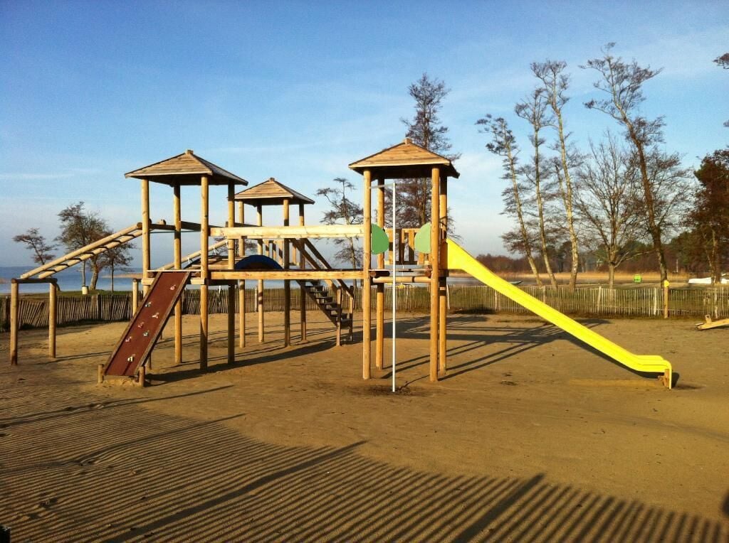 A wooden playground structure with slides and climbing elements, surrounded by sand and trees.