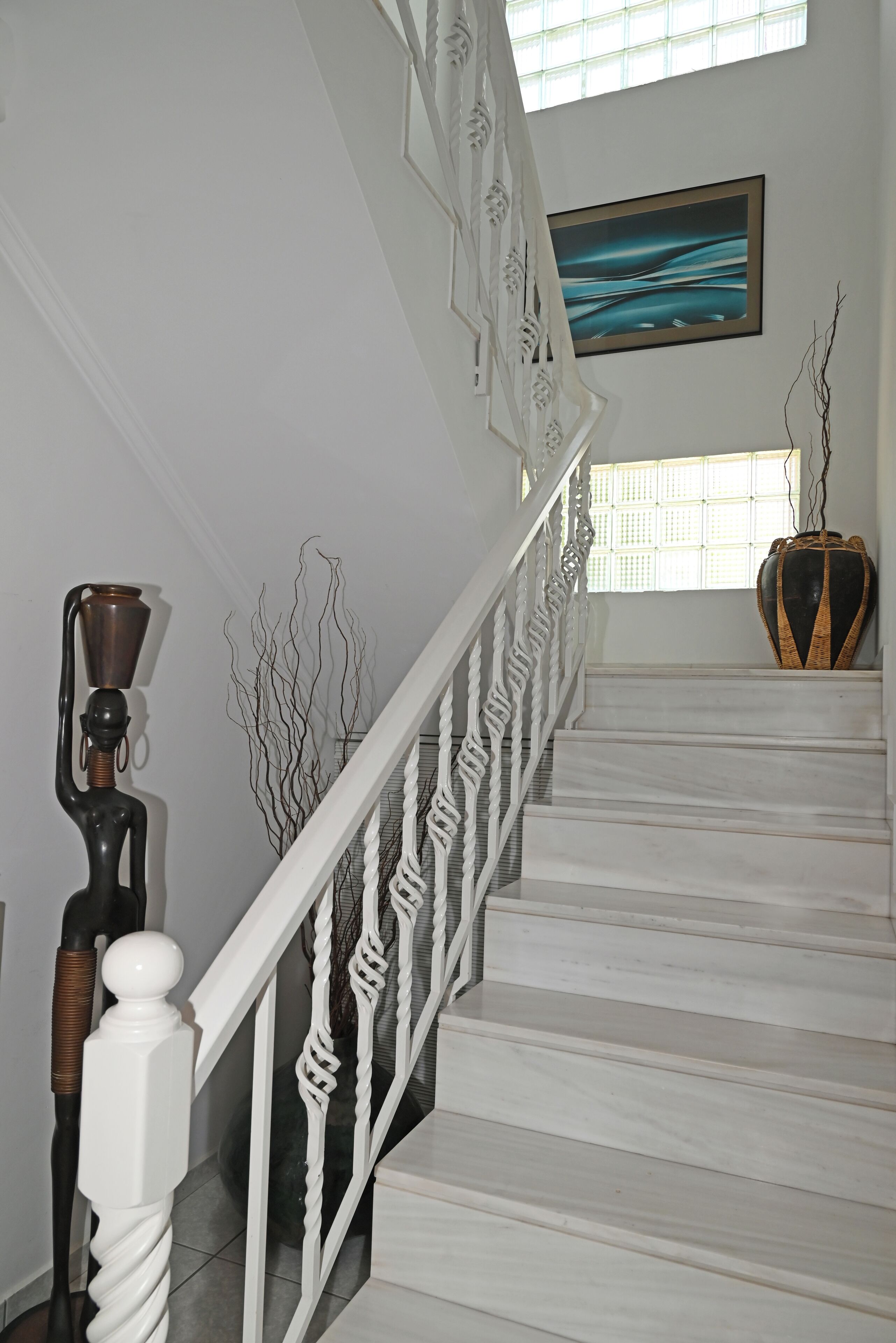 A staircase with white railings and a marble floor, leading to a room with a large window and a framed abstract painting.