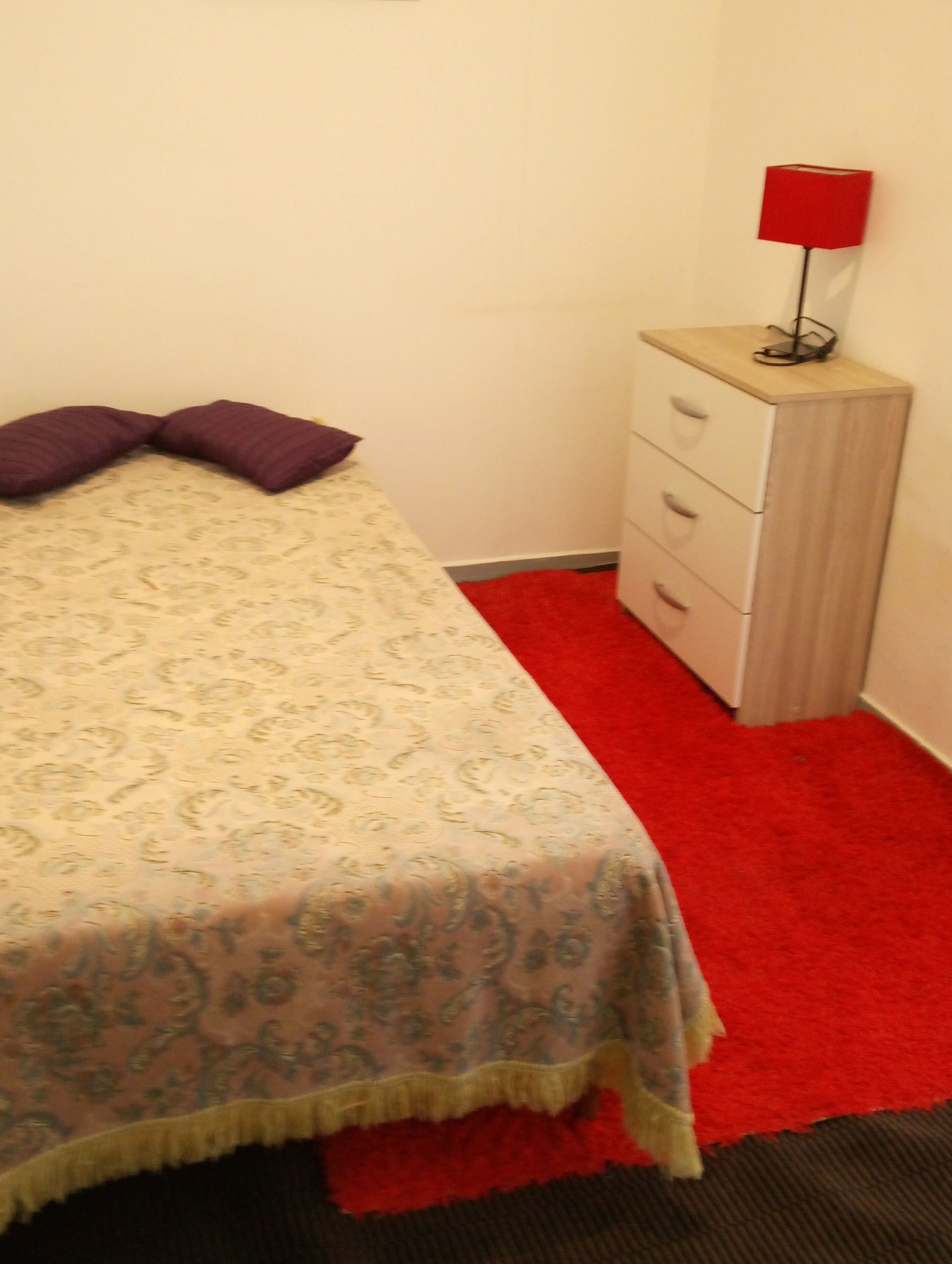 A small room with a bed, a red carpet, a white dresser, and a red lamp.