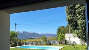 Vista dalla finestra su un giardino con piscina, prato verde e montagne in lontananza.