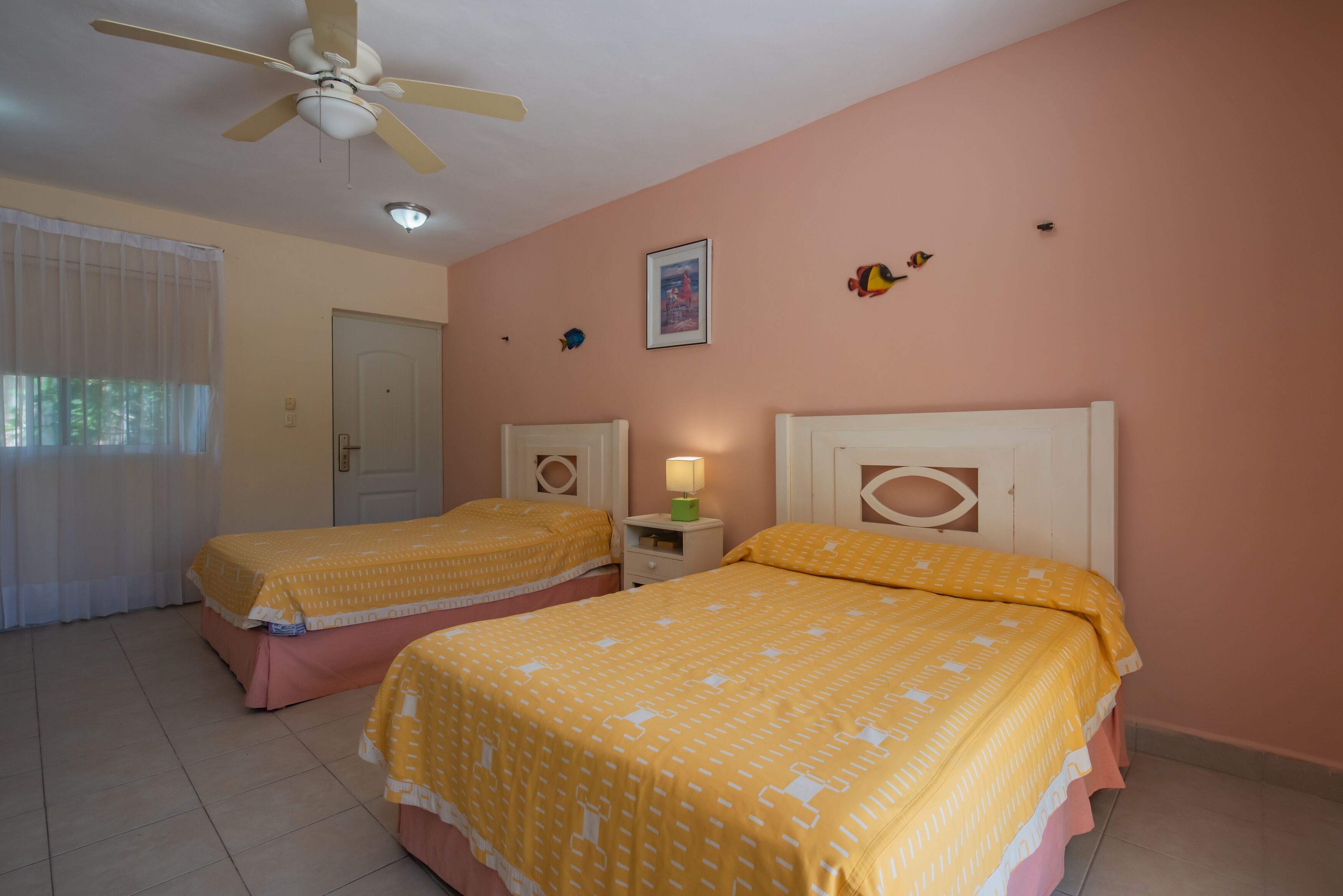 A bedroom with two beds, a ceiling fan, a nightstand with a lamp, and a door.