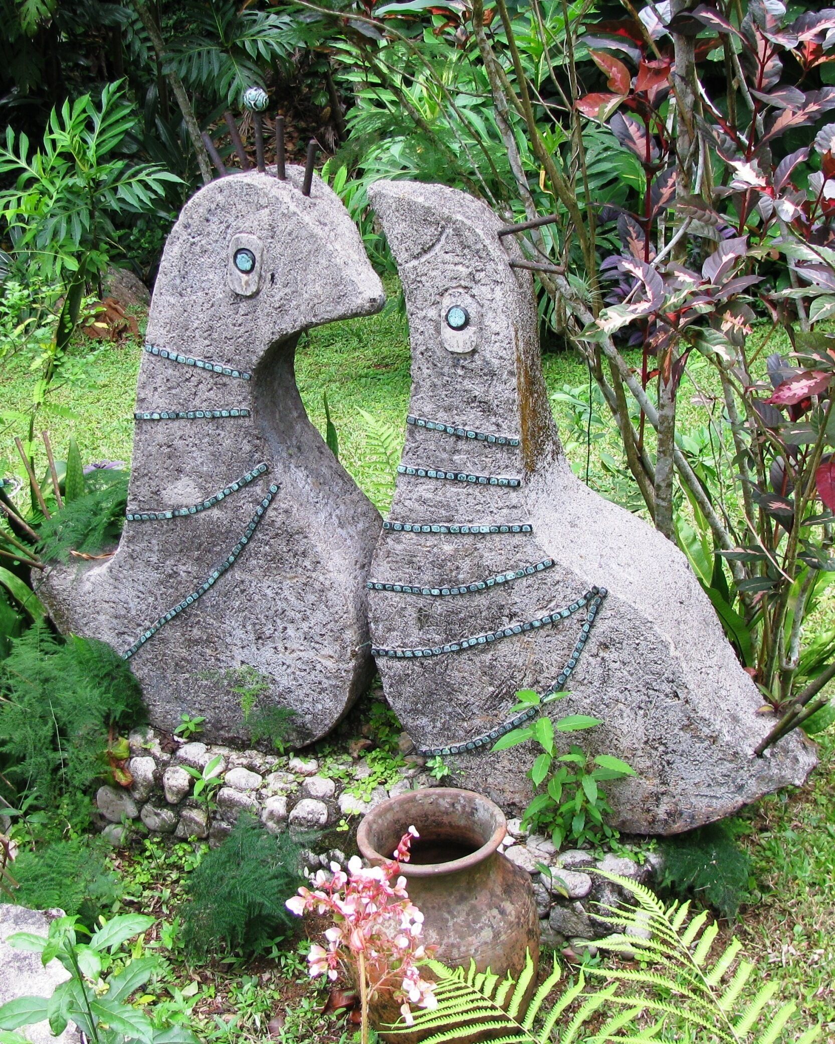Two stone sculptures of birds with chains, surrounded by lush greenery and a small pot.