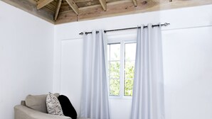 A room with a beige sofa, a window with white curtains, and a wooden ceiling. - Pulse Suites at Villa Ronai (Kingston)