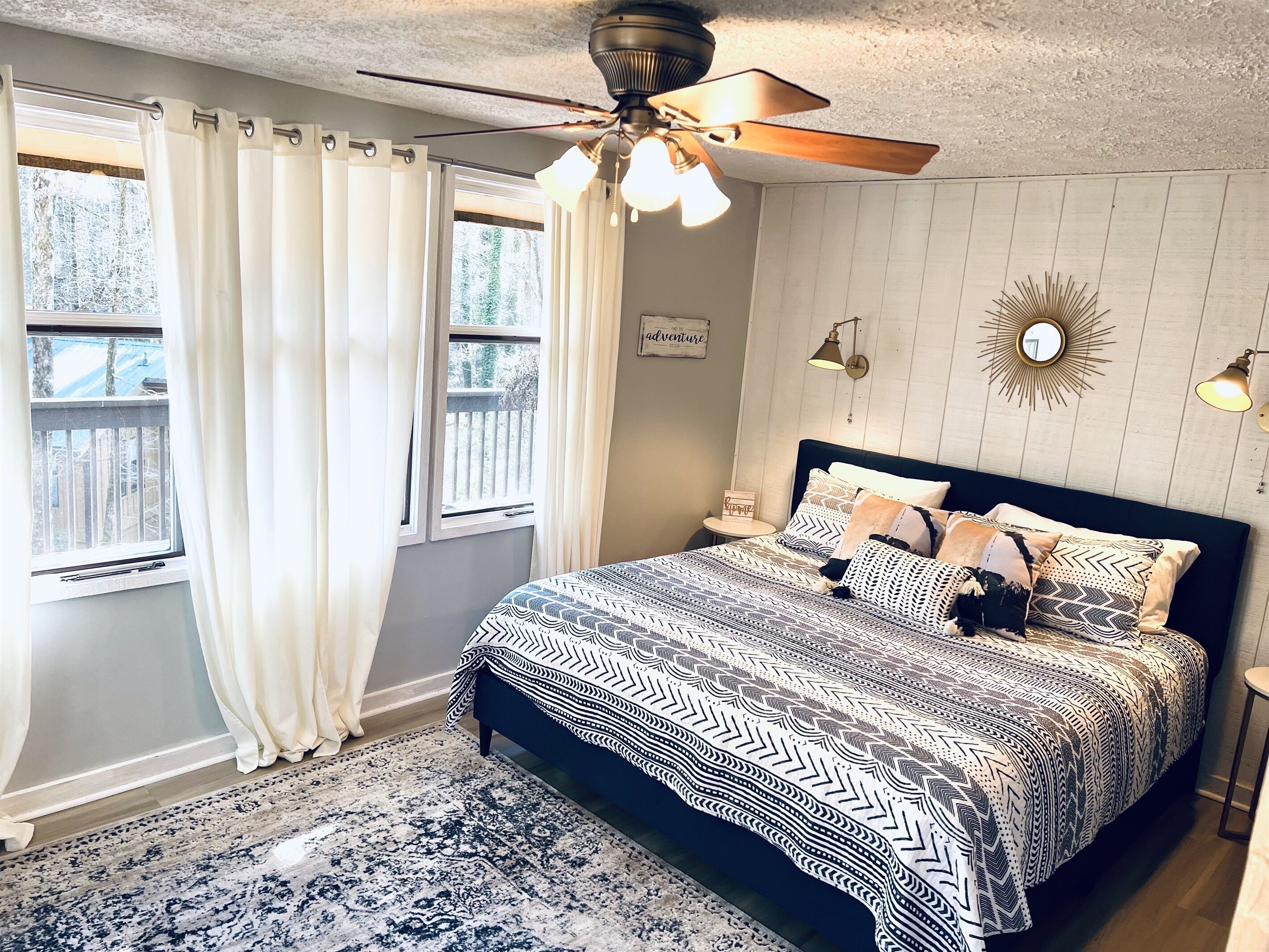 A bedroom with a bed, a ceiling fan, a window with curtains, a small side table, and a wall-mounted mirror.