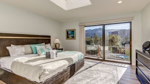 A bedroom with a bed, a nightstand, a lamp, a painting, and a view of mountains through a sliding glass door.