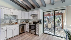 A modern kitchen with white cabinetry, a stainless steel dishwasher, and a built-in oven. There is a large sliding glass door leading to an outdoor patio area with a dining table and chairs.