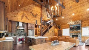 L’intérieur d’un chalet rustique, avec une cheminée en pierre, des poutres en bois, une cuisine avec un mur de fond en pierre et un escalier menant à un étage supérieur.