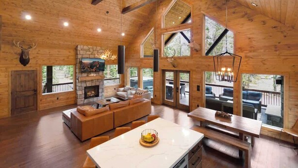 Un intérieur spacieux de chalet en bois, avec une cheminée, un grand canapé, une table à manger et une vue sur l’extérieur.