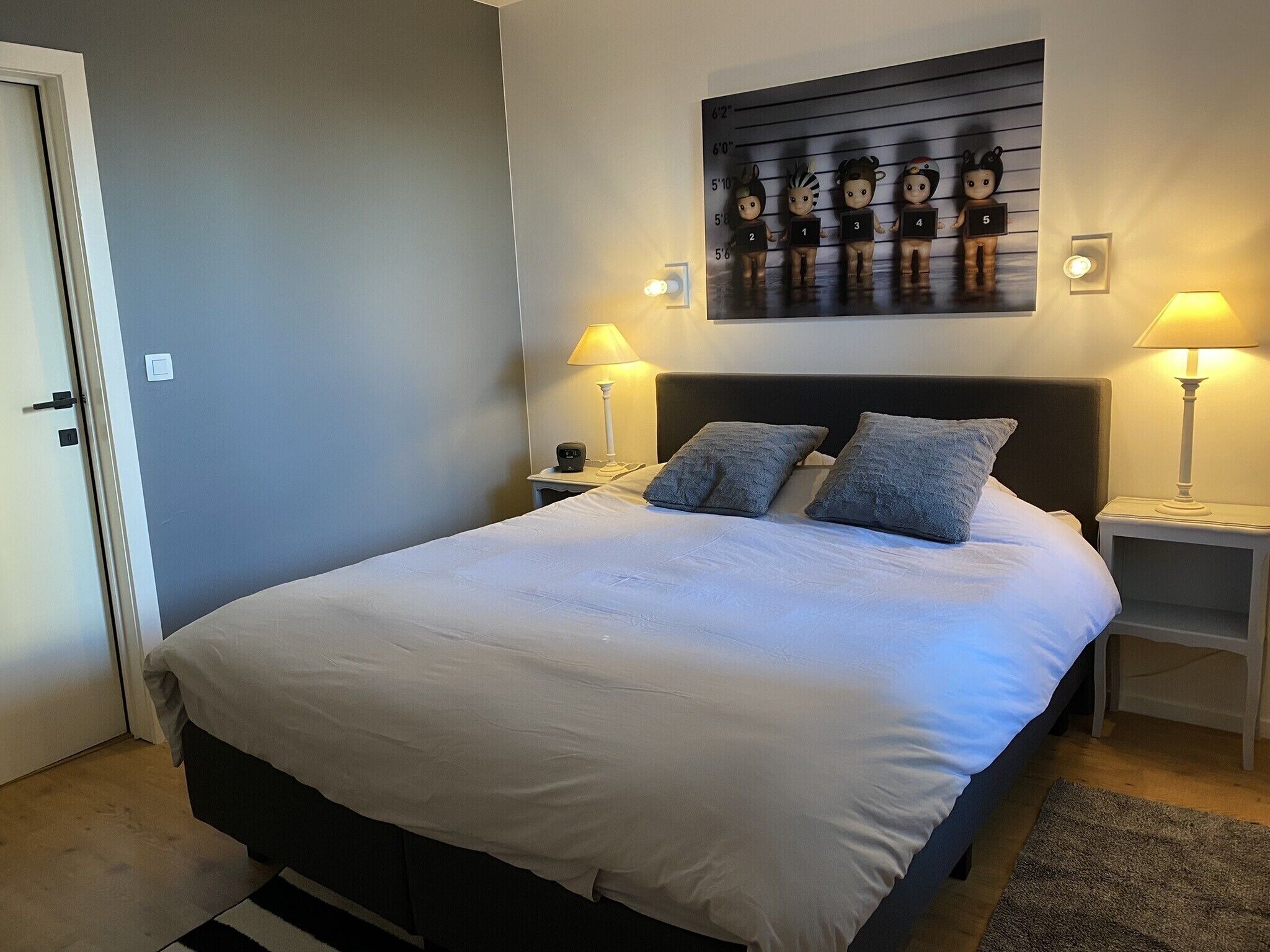 A neatly made bed with white linens and two pillows, flanked by bedside tables with lamps, a wall-mounted artwork, and a door to the left.