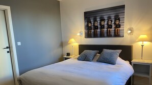 A neatly made bed with white linens and two pillows, flanked by bedside tables with lamps, a wall-mounted artwork, and a door to the left. - NICE TWO-BEDROOM APPARTMENT WITH TERRACE (Oostende)