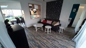 A modern living room with a sofa, two round tables, and a coffee table. There is a kitchen area with a counter and chairs visible in the background. - LA GABARDISE (Plouarzel)