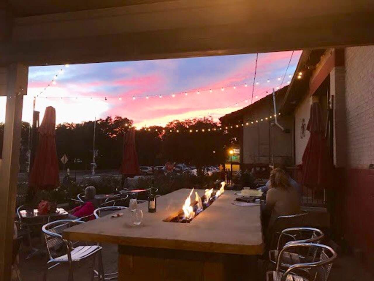 Un espace extérieur aménagé pour les repas, avec des tables et des chaises, un foyer et des guirlandes lumineuses, le tout sur fond de ciel au coucher du soleil.