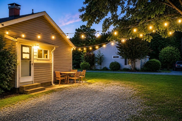 Une maison avec une allée en gravier, une table et des chaises en bois à l’extérieur, et des guirlandes lumineuses.