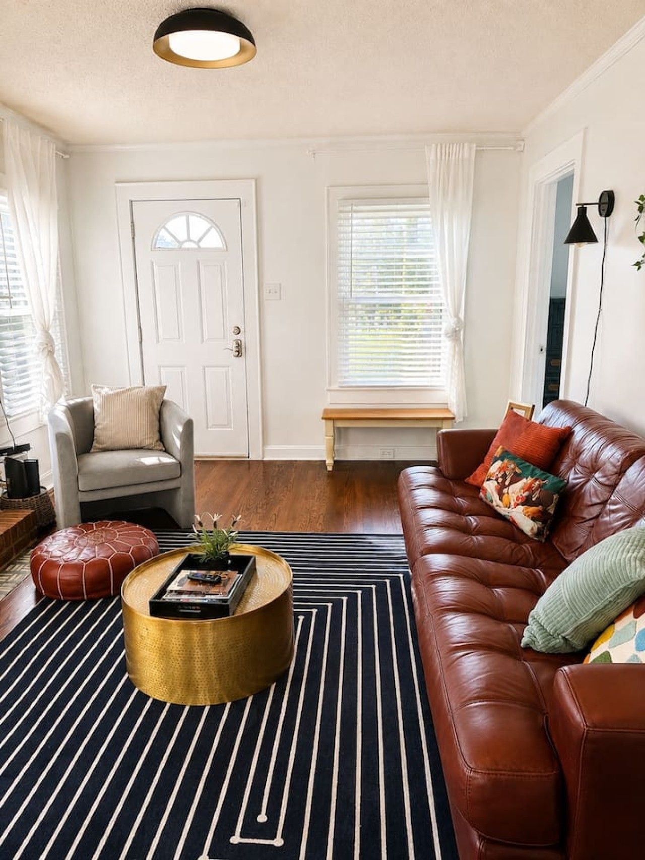 A living room with a brown leather sofa, a round coffee table, a striped rug, a white door, and a window with blinds.