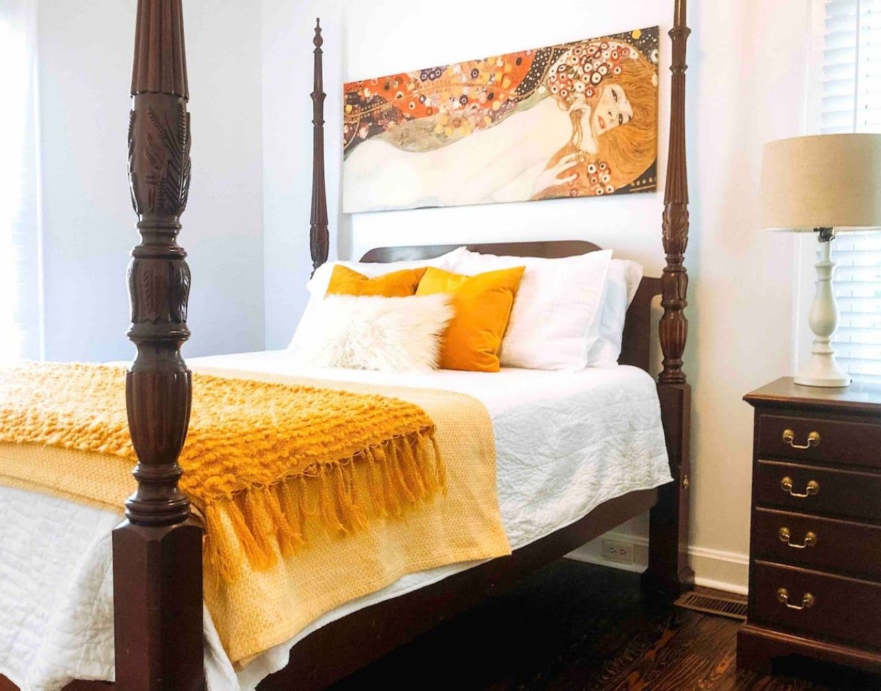 Un letto a baldacchino con copriletto bianco e giallo, un comodino in legno con cassetti e un quadro sopra il letto.