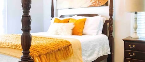 Un letto a baldacchino con copriletto bianco e giallo, un comodino in legno con cassetti e un quadro sopra il letto.