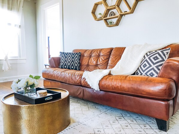 Un sofá de cuero marrón con cojines estampados, una mesa de centro tejida y un adorno hexagonal de madera en la pared.