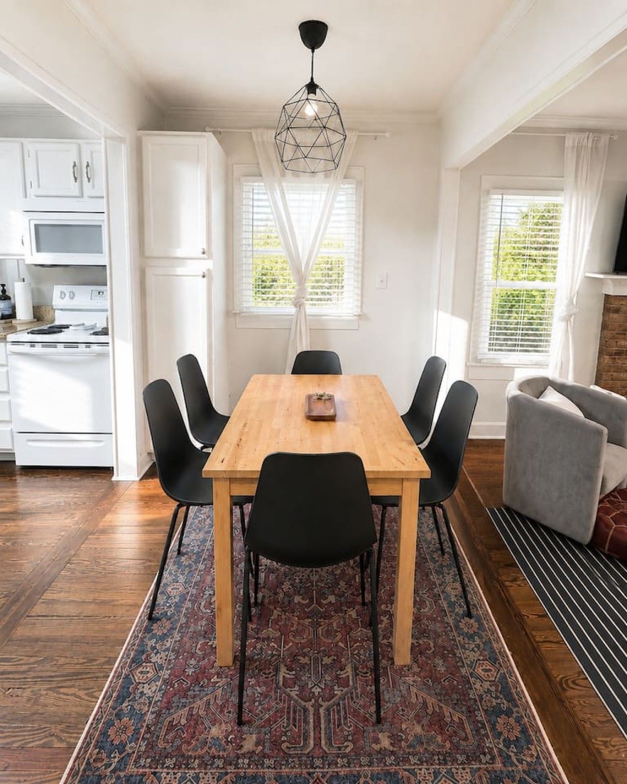 A dining area with a wooden table, black chairs, a patterned rug, and a kitchen in the background.
