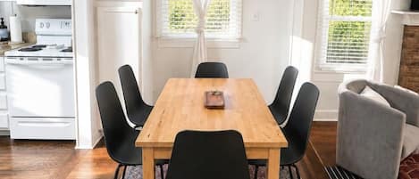 A dining area with a wooden table, black chairs, a patterned rug, and a kitchen in the background.