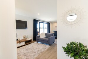 A modern living room with a grey sofa, a flat-screen TV mounted on the wall, a wooden side table, and a large mirror with a sunburst design. - Tussock Treasure 1 - Twizel (Twizel)