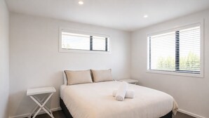Une chambre à coucher avec un lit, deux tables de chevet et deux fenêtres équipées de stores.
