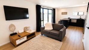 Un salon moderne avec un téléviseur à écran plat, une table basse en bois, un canapé gris et un coin cuisine à l’arrière-plan.