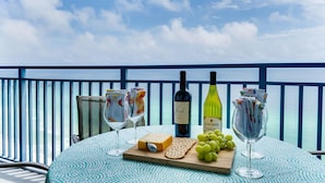A table with a bottle of wine, cheese, crackers, and grapes on a balcony with a view of the sea.