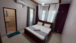 A bedroom with a wooden bed, a bedside table, a window with sheer curtains, and an air conditioning unit on the wall.