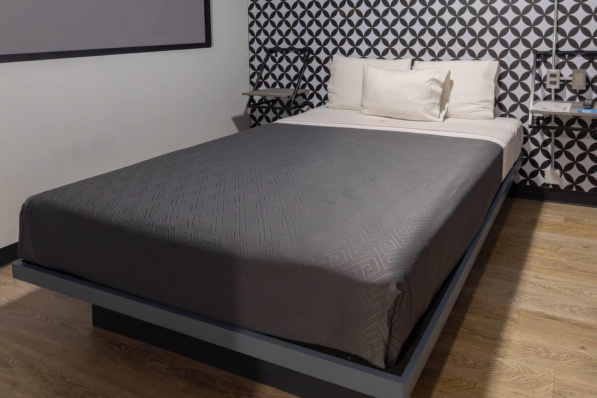 A bed with a patterned bedspread, a bedside table with a lamp, and a wall with a geometric pattern.
