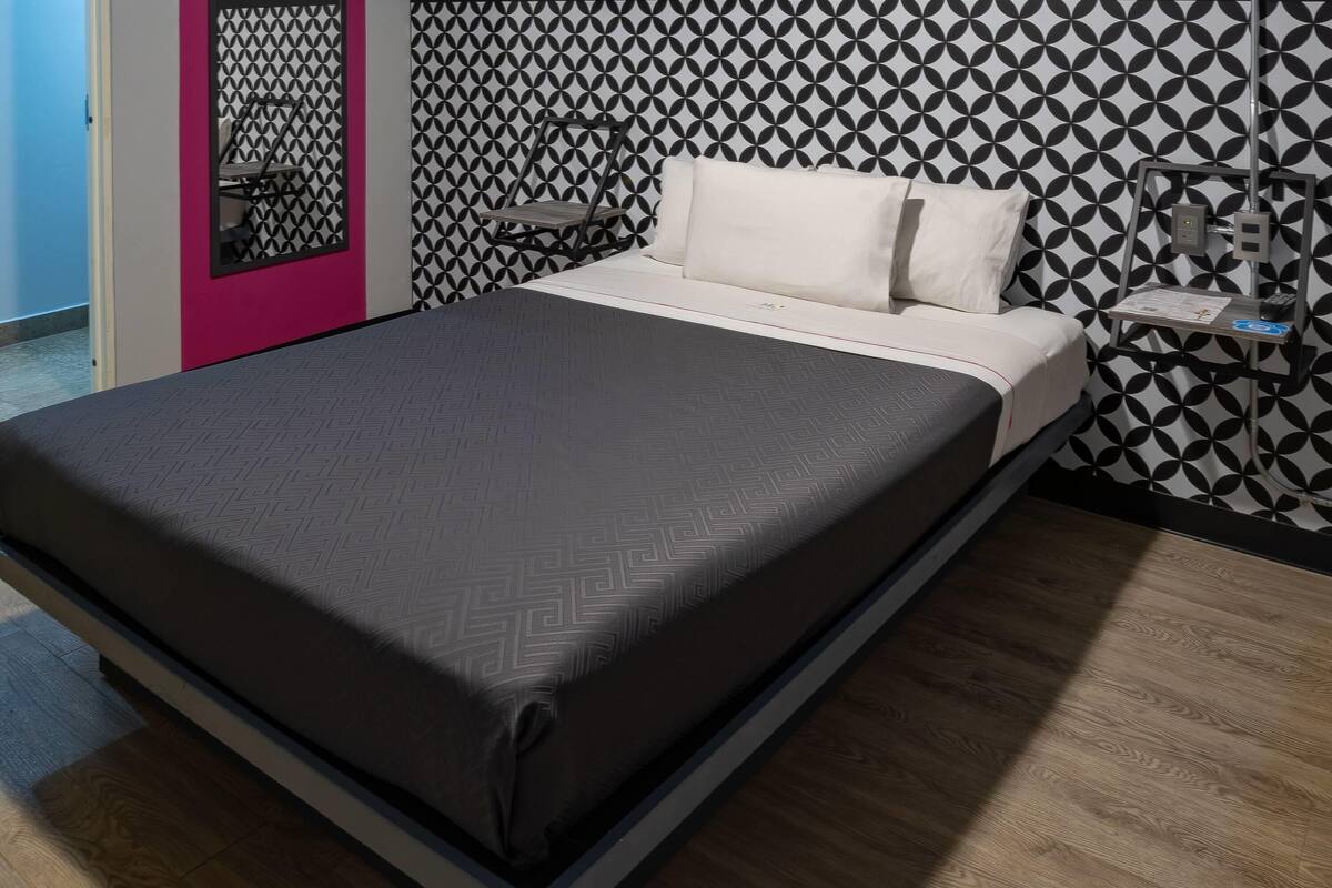 Ein Bett mit gemustertem Überwurf, ein Nachttisch mit Lampe sowie eine Wand mit geometrischem Muster.