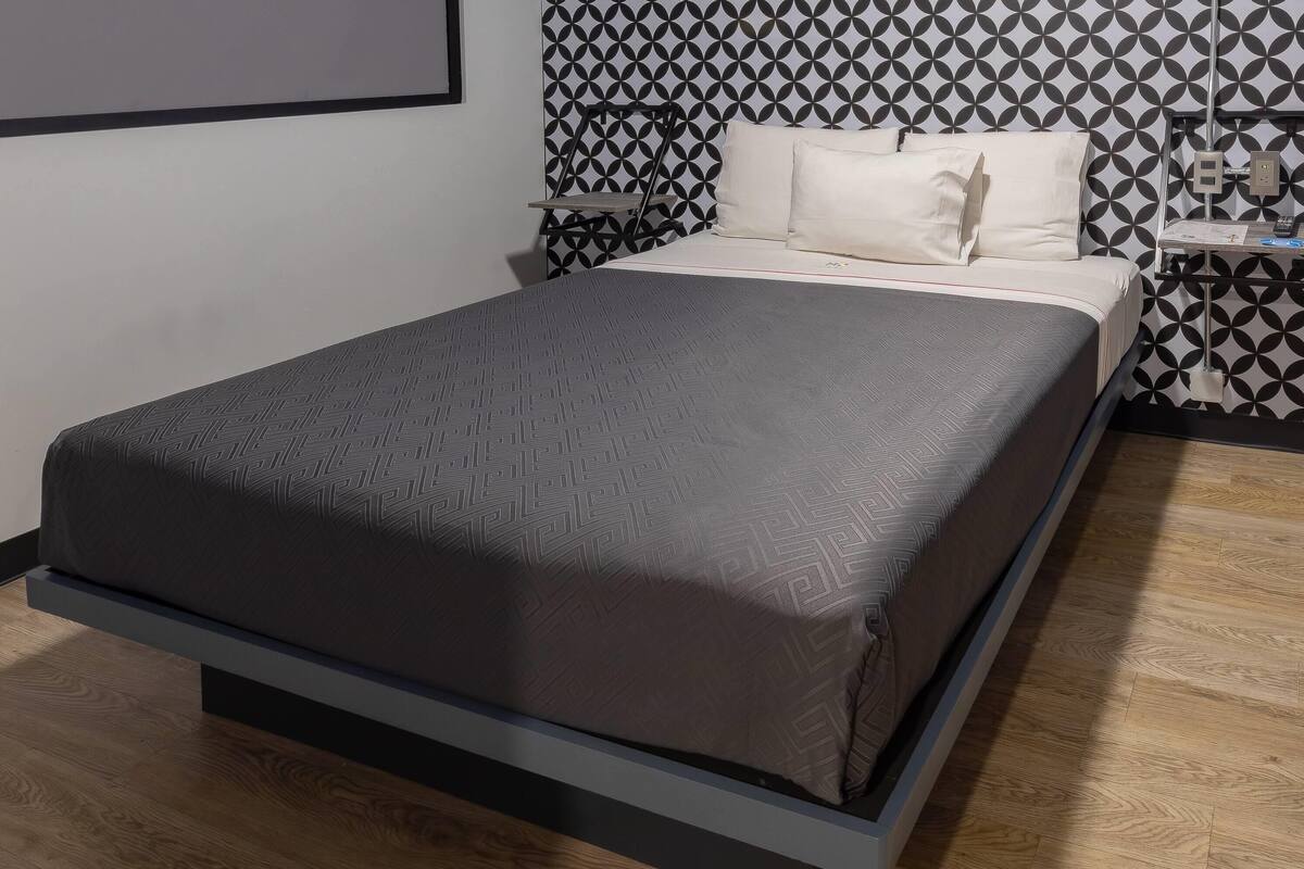 Ein Bett mit gemustertem Bettbezug, ein Nachttisch mit Lampe und eine Wand mit geometrischem Muster.