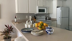 A kitchen with white cabinets, a microwave, a refrigerator, and a fruit bowl on the counter. - Sun Filled Townhome w/Pool, 5' to beach, walk to bars/restos, luxury bath/shower (Marco Island)