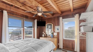 Une chambre à coucher avec un grand lit, des poutres apparentes en bois au plafond, un ventilateur de plafond, une commode, un téléviseur et de grandes fenêtres avec des rideaux.