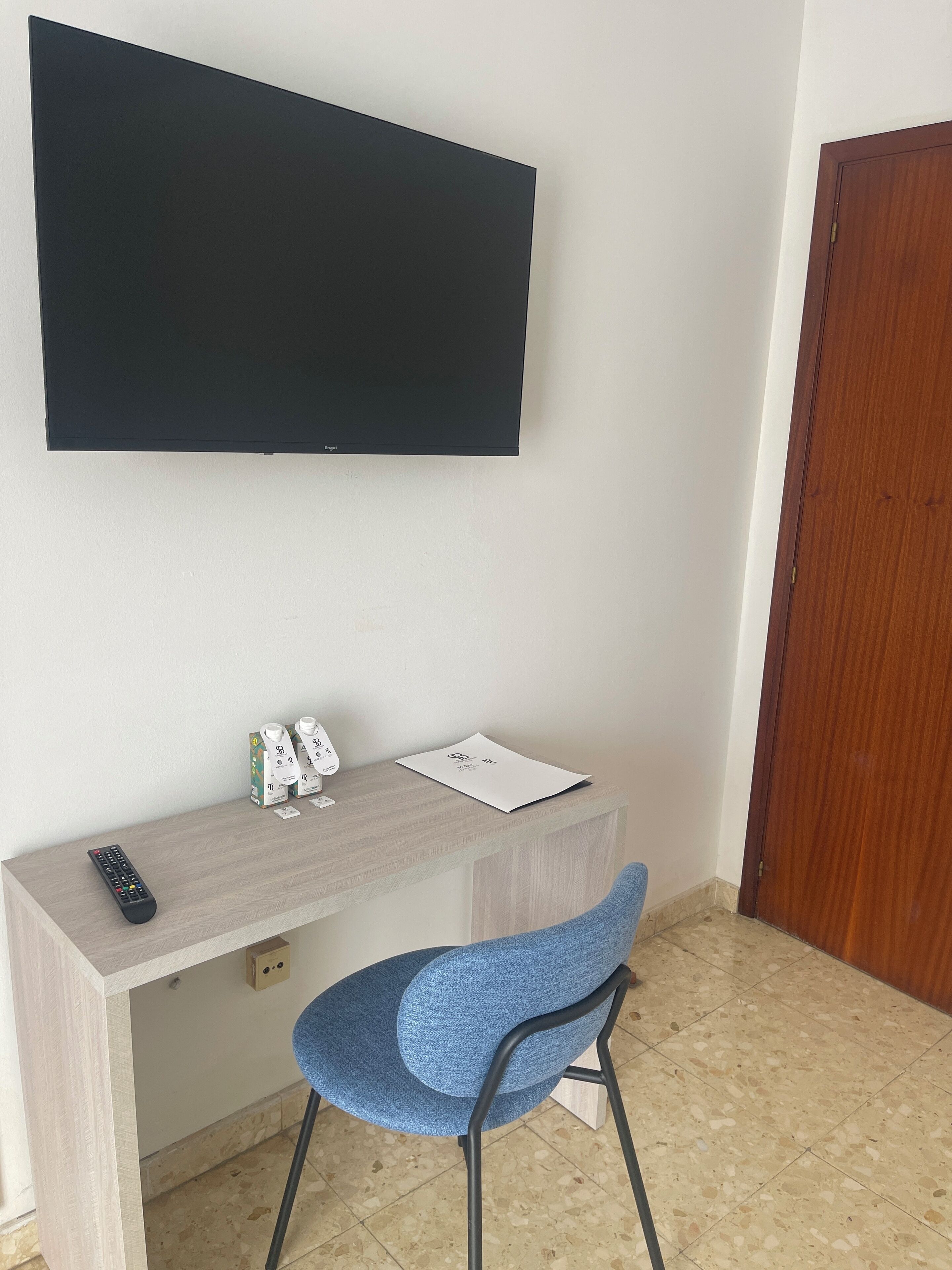 A room with a flat-screen TV mounted on the wall, a wooden desk with a chair, a remote control, and some items on the desk.