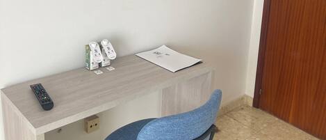 A room with a flat-screen TV mounted on the wall, a wooden desk with a chair, a remote control, and some items on the desk.