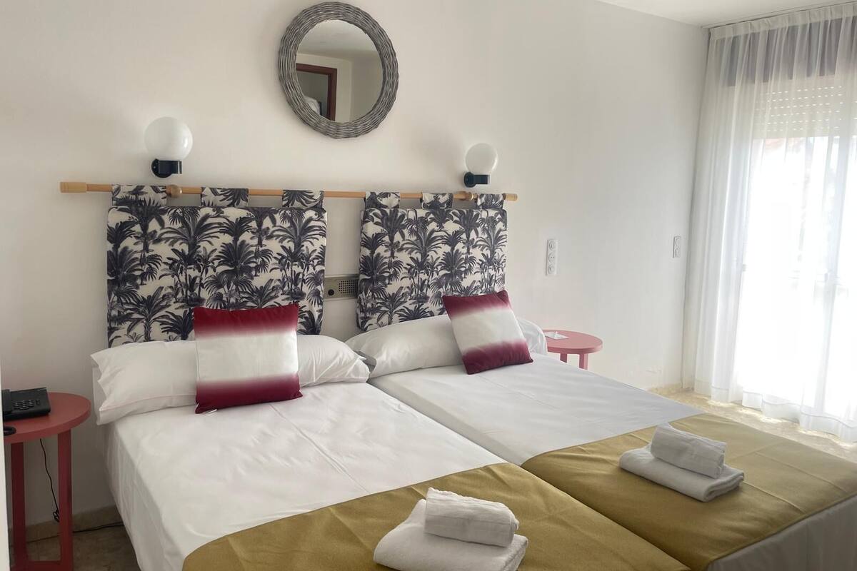 A hotel room with two beds, a round mirror, a small table, and a window with sheer curtains.