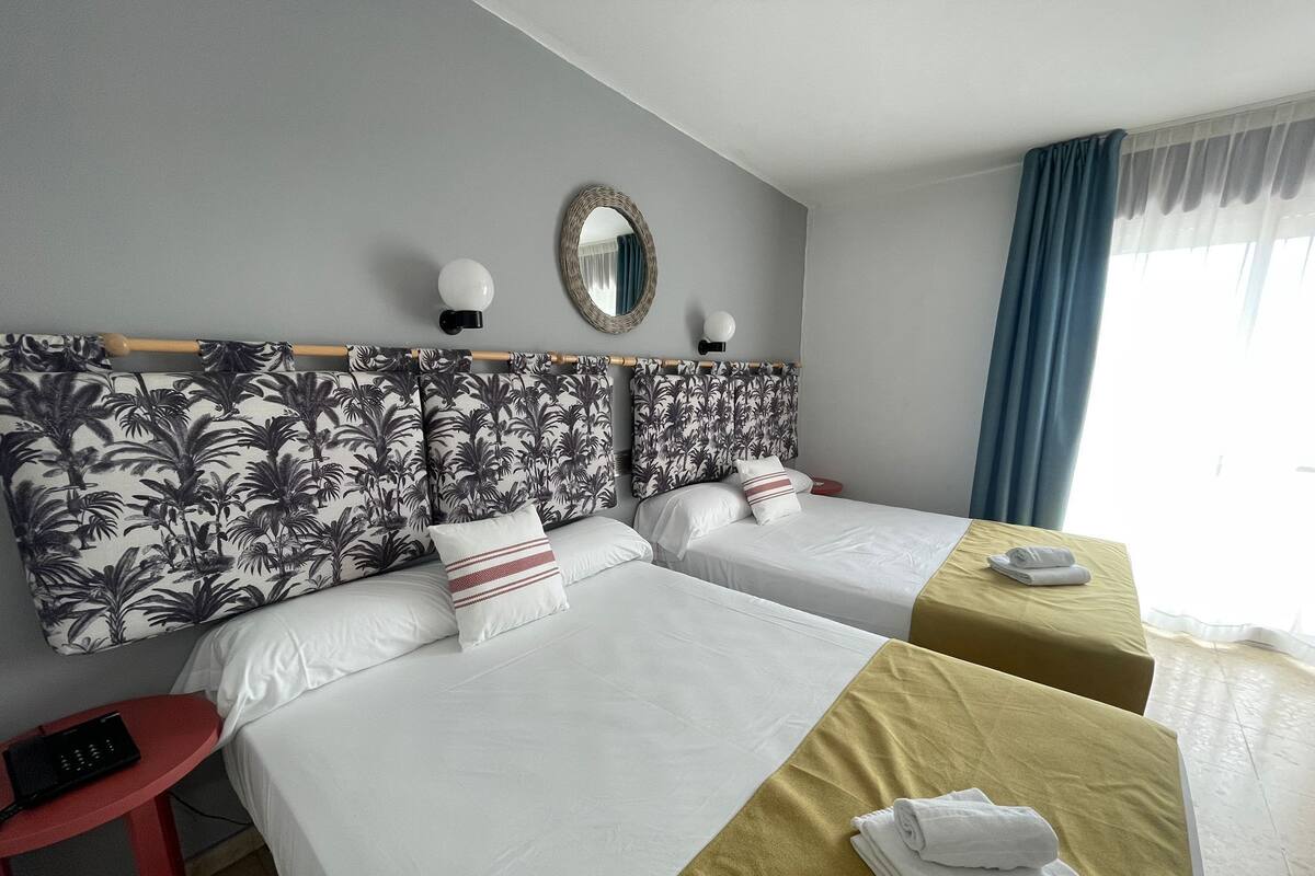 A hotel room with two beds, a red side table, a mirror, and a window with blue curtains.