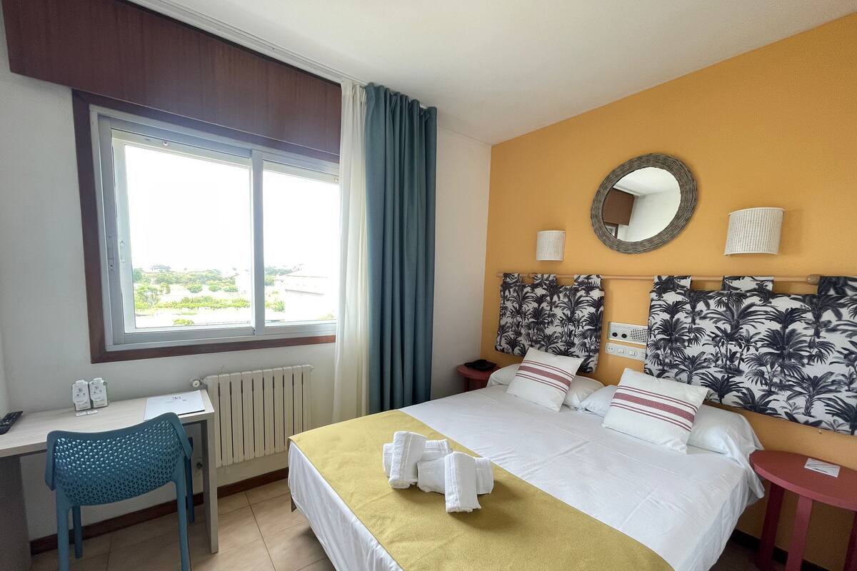 A hotel room with a bed, a desk, a chair, a small table, a window with curtains, and a wall with a mirror and patterned wallpaper.
