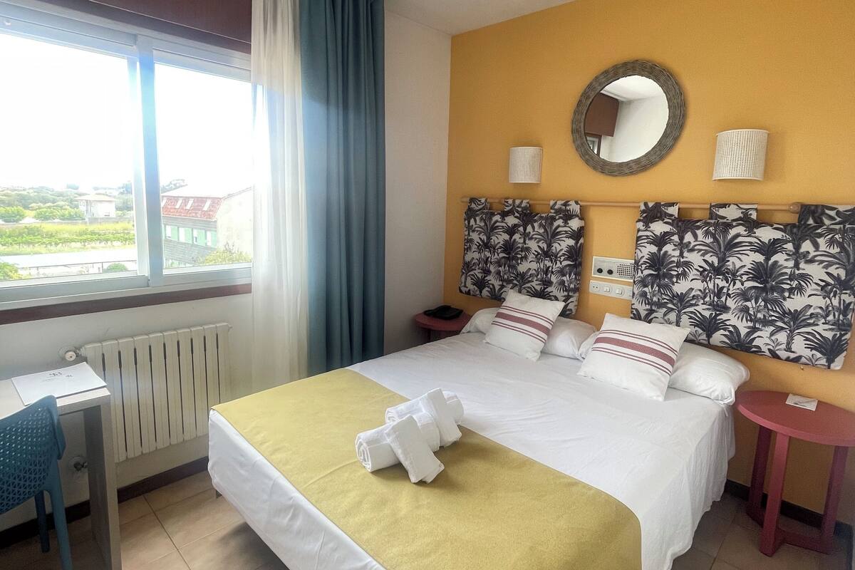 A hotel room with a bed, a desk, a chair, a small table, a window with curtains, a radiator, and a view of the outside.