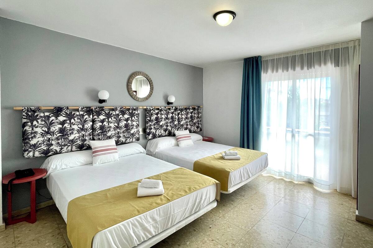 A hotel room with two beds, a mirror, a small table, and a window with curtains.