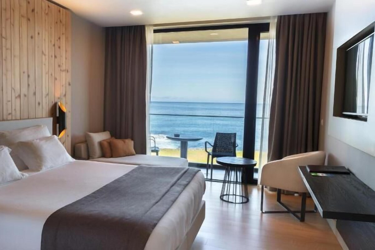 Ein Hotelzimmer mit einem großen Bett, einem Schreibtisch, einem Sessel und Blick auf das Meer.