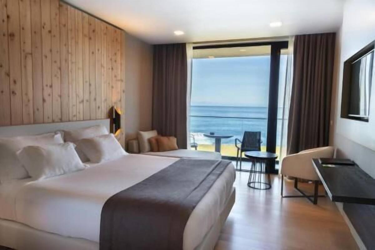 Ein Hotelzimmer mit einem großen Bett, einem Schreibtisch, einem Sessel und Blick aufs Meer.