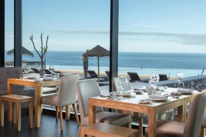Un espace repas avec des tables disposées pour accueillir quatre personnes, donnant sur une plage et sur une structure recouverte de chaume.