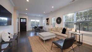 Un vaste salon comprenant un grand canapé, une table basse et un téléviseur fixé au mur. On y trouve également un coin repas avec une table et des chaises, ainsi qu’une cuisine à l’arrière-plan.