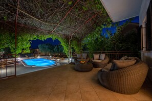 Un espace extérieur avec une piscine, des meubles en osier et une vue sur les arbres la nuit.