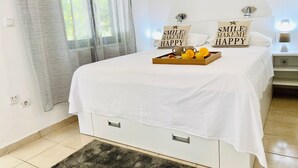 Un lit bien fait, avec un plateau contenant du jus d’orange et un oreiller orné du texte « SMILE MAKEME HAPPY », un mur blanc, une fenêtre avec des rideaux légers et une table de chevet avec une lampe.