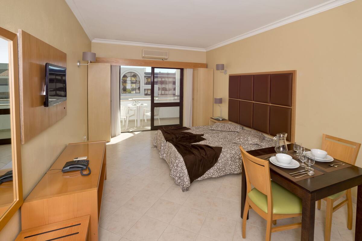 Quarto de hotel com cama, escrivaninha com telefone, mesa de jantar posta para dois e TV na parede.