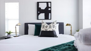A neatly arranged bedroom with a bed, pillows, bedside lamps, a framed artwork on the wall, and a window with blinds.