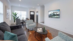 A modern living room with a sofa, armchairs, a coffee table, and a dining area with a table and chairs. There is a framed picture on the wall and a cityscape poster.