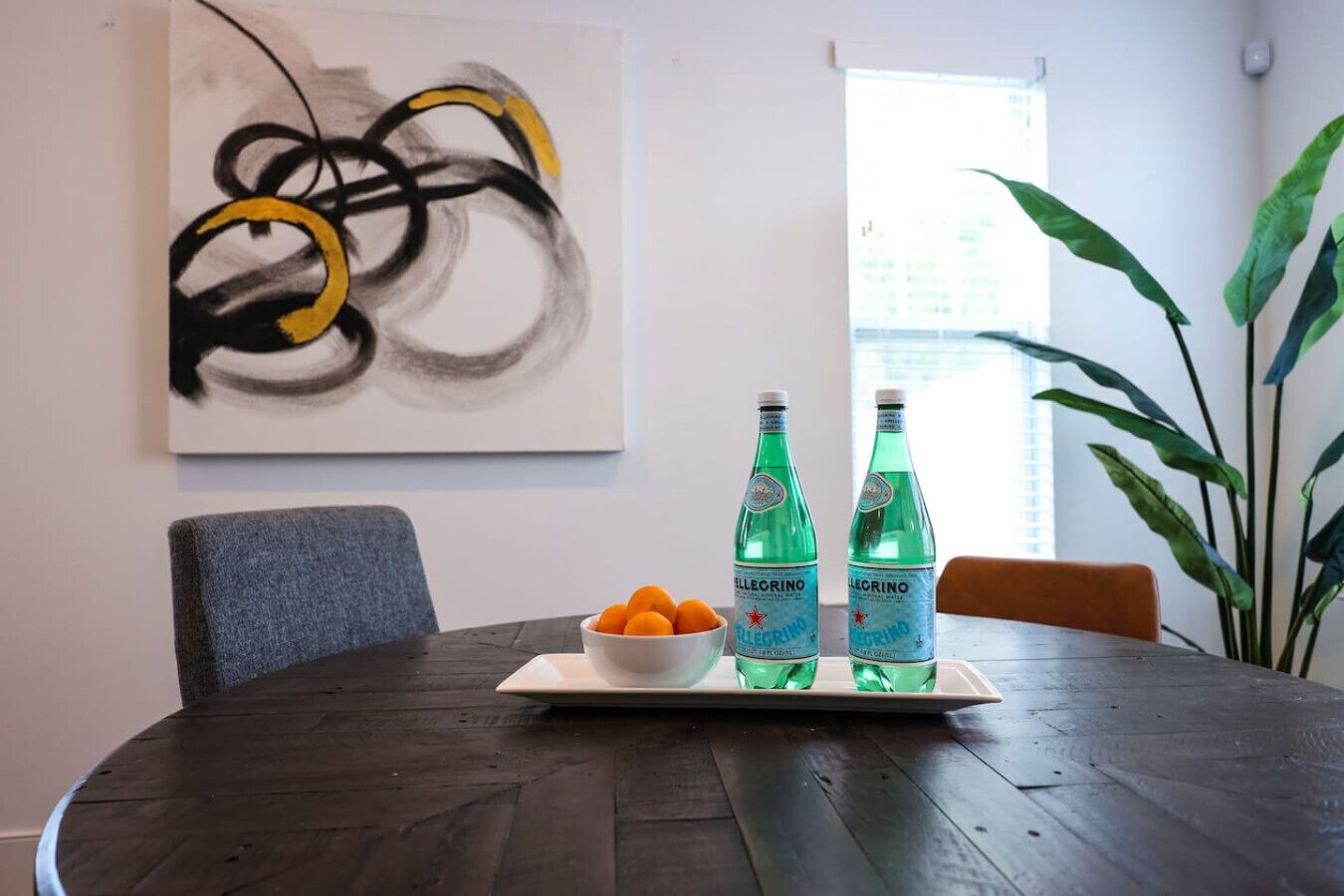 A dining table with two bottles of Pellegrino water, a bowl of oranges, and a modern abstract painting on the wall.